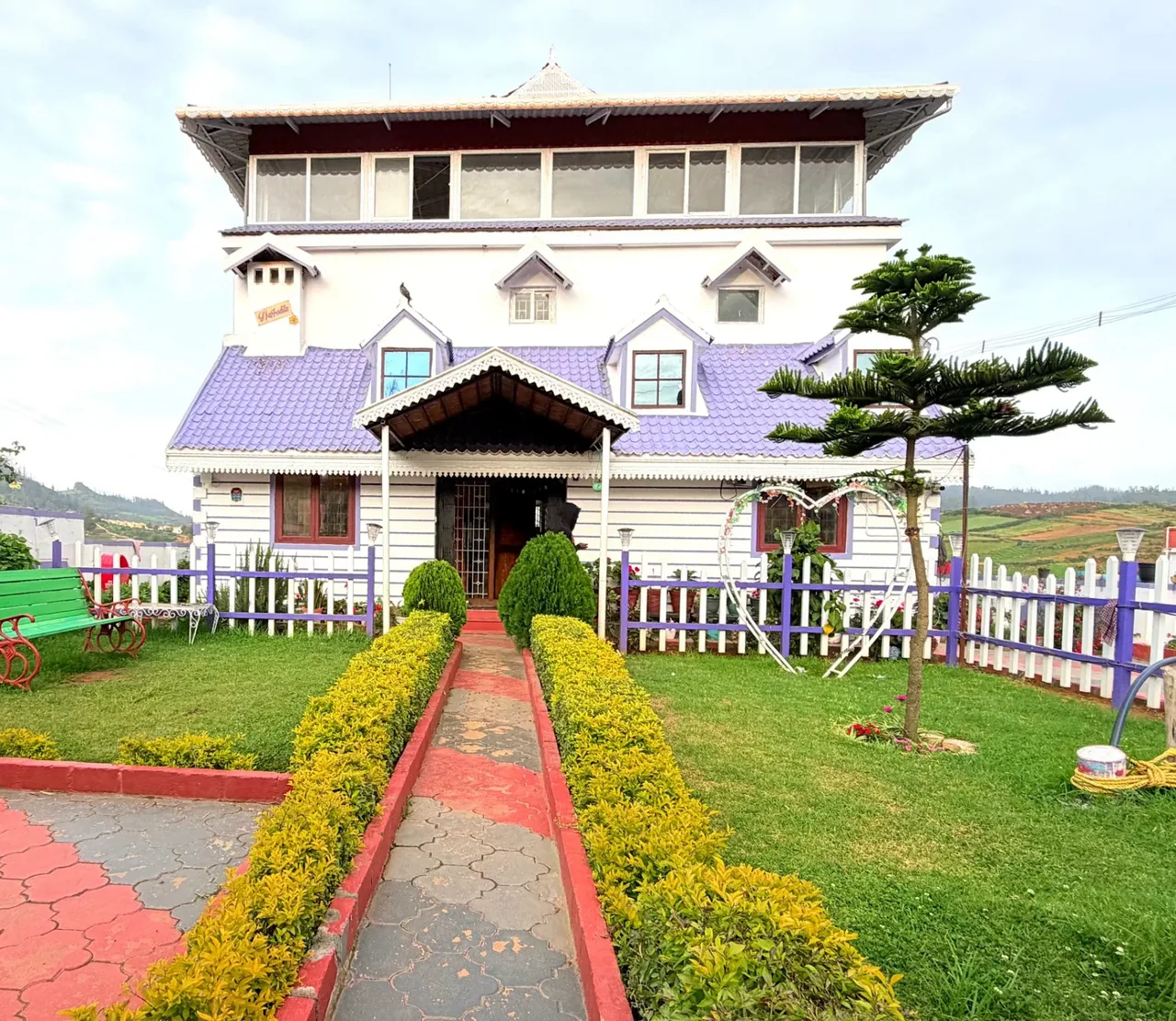 The 5 BHK villa: a white three-storey house with a lilac roof, a red garden path lined with yellow hedges, and lawns on both sides